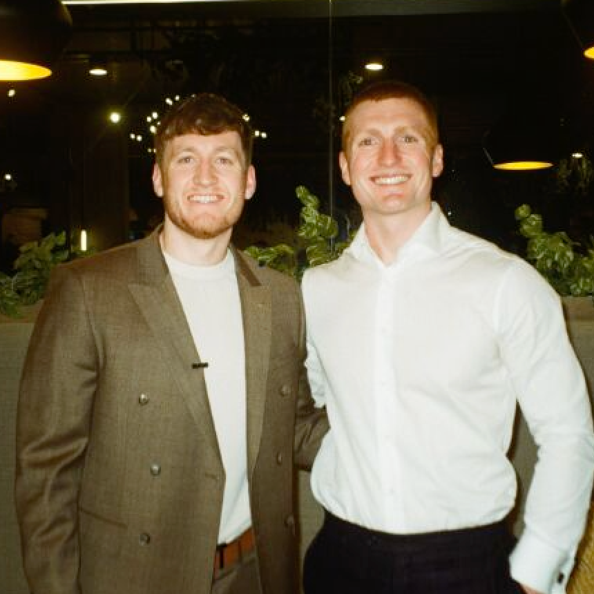Two smiling men standing side by side indoors, wearing a brown jacket and white shirt
