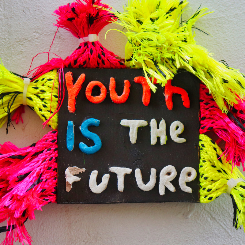 Black sign reading Youth is the future in colorful letters surrounded by neon pink and yellow tassels