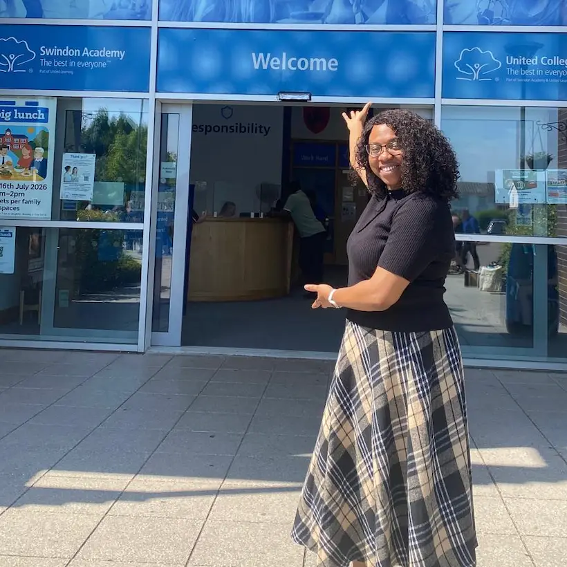 A smiling woman in a plaid skirt gestures toward the entrance of an academy building