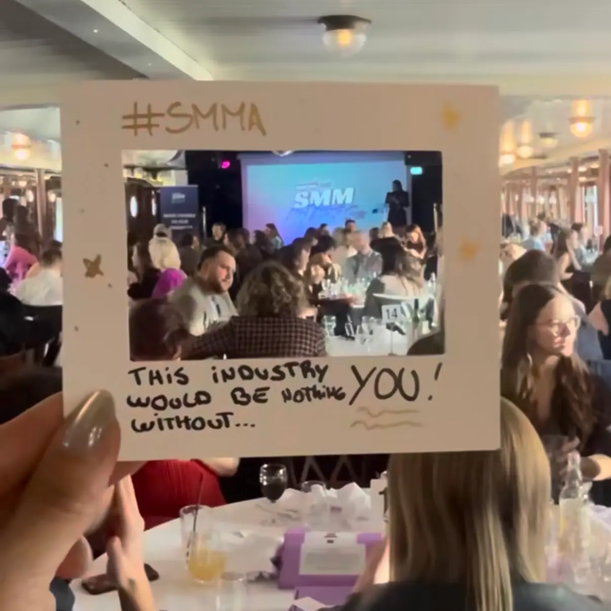 A hand holds a paper photo frame cutout over a crowded awards event with attendees at tables